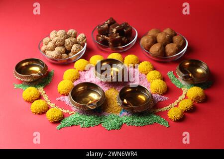 Happy Diwali. Diya-Lampen, farbenfrohe Rangoli, Blumen und köstliche indische Süßigkeiten auf rotem Tisch Stockfoto