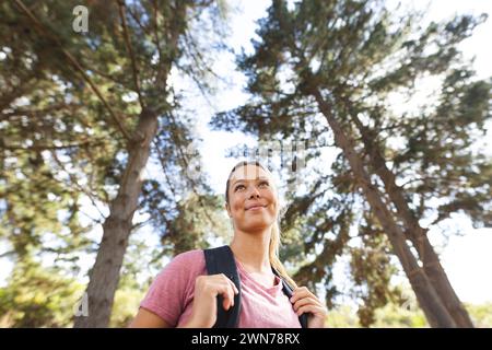 Junge kaukasische Frau mit Rucksack schaut nachdenklich zwischen hohen Bäumen auf einer Wanderung auf Stockfoto