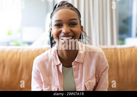 Eine junge Afroamerikanerin lächelt hell, sitzt auf einem braunen Sofa auf einem Videoanruf Stockfoto