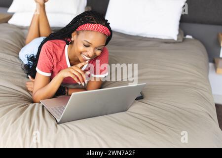 Die junge afroamerikanische Frau liegt auf ihrem Bauch mit einem Laptop auf einem Bett Stockfoto