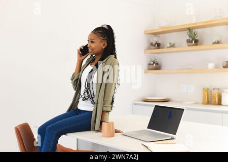 Die junge afroamerikanische Frau plaudert am Telefon, sitzt neben einem Laptop in einer modernen Küche Stockfoto