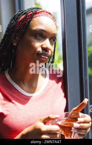 Eine junge Afroamerikanerin schaut aus dem Fenster und hält eine Tasse Tee Stockfoto