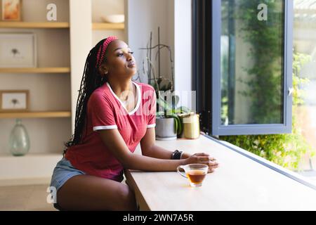 Eine junge Afroamerikanerin schaut aus einem Fenster, eine Tasse Tee vor sich, mit Kopierraum Stockfoto