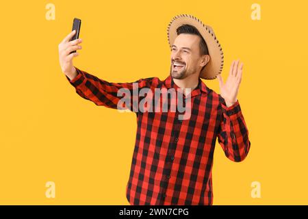 Ein gutaussehender Mann mit Handy, der Selfie auf gelbem Hintergrund macht. Festa Junina (Juni-Festival) Stockfoto