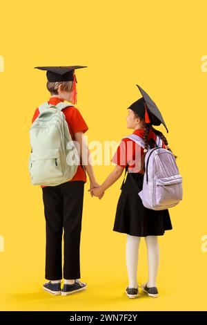 Kleine Kinder in Graduiertenhüten auf gelbem Hintergrund, Rückansicht. Ende des Schuljahres Stockfoto