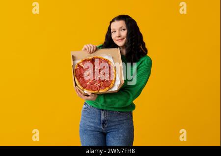 Schöne junge Frau, die Pappschachtel mit leckerer Pizza auf gelbem Hintergrund hält Stockfoto