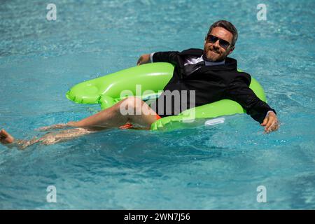 Geschäftsmann im Anzug mit Laptop im Swimmingpool. Verrückter Geschäftsmann schwimmt im Sommerurlaub. Aufgeregter Geschäftsmann in Neoprenanzug im Swimmingpool Stockfoto