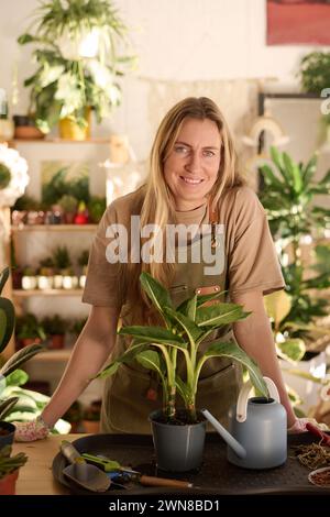Porträt einer modernen kaukasischen Frau mit Schürze, die am Tisch in der Werkstätte steht und in die Kamera lächelt Stockfoto