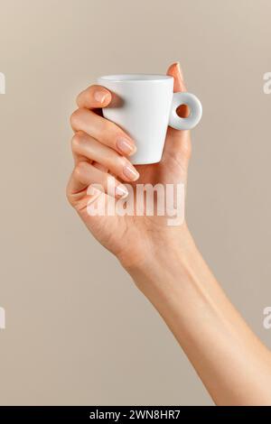 Anonyme Frau, die keramische weiße Mini-Kaffeetasse mit Griffring in der gepflegten schlanken Fingerhand gegen beigefarbenen Hintergrund im Licht trägt Stockfoto