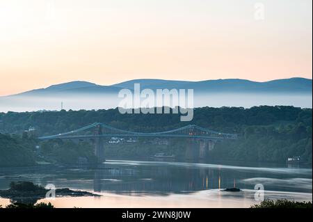 Die Menai Bridge, die von Nordwales zur Insel Angelsey führt, spiegelt sich im Wasser wider. Es ist Morgengrauen und eine Schicht niedriger Wolken ist zu sehen Stockfoto