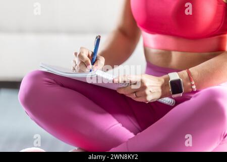 Sportliche Frau sitzt auf einer Matte und schreibt zukünftige Trainingspläne auf, um sportliche Ergebnisse zu erzielen. Stockfoto