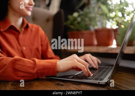 Gärtnerin sitzt auf einem Sessel im Gewächshaus, benutzt Laptop und spricht im Internet in der Nähe von Zimmerpflanzen. Freiberufliches Konzept Stockfoto