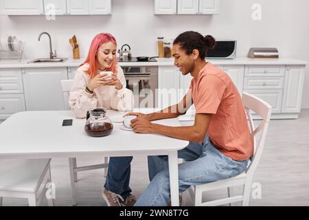 Gut aussehendes, fröhliches, abwechslungsreiches Paar, das am Tisch mit Kaffee sitzt und sich ansieht Stockfoto