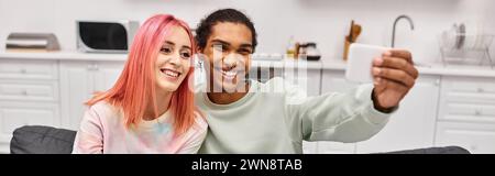Fröhliches attraktives multikulturelles Paar, das Selfies im Wohnzimmer zu Hause nimmt, Banner Stockfoto