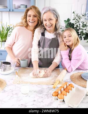 Zärtlicher Moment, in dem Großmutter, Mutter und Enkelin Teig auf Küchentisch mit Eiern, Mehl und Milch zusammenkneten. Familienporträt in der Küche Stockfoto
