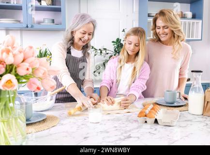 Glückliche Seniorengroßmutter, die der süßen Enkelin mit ihrer Mutter beibringt, Teig zu Rollen, um selbstgemachte Kekse zu kochen. Familie genießt es, Zeit miteinander zu verbringen Stockfoto