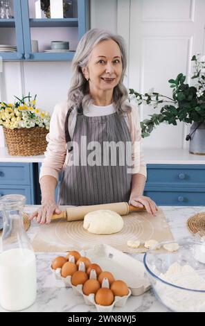 Frauen der älteren Generation. Grauhaarige Großmutter, die Köstlichkeiten aus Teig mit sanften Händen zubereitet, ihre Küche ist voller Liebe und Traditionen Stockfoto