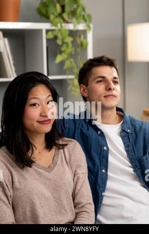 Lächelnder junger Erwachsener vielfältiges Paar, das sich zu Hause auf dem Sofa entspannt. Frau, die in die Kamera schaut, neben einem glücklichen Mann, der auf einer Couch sitzt Stockfoto