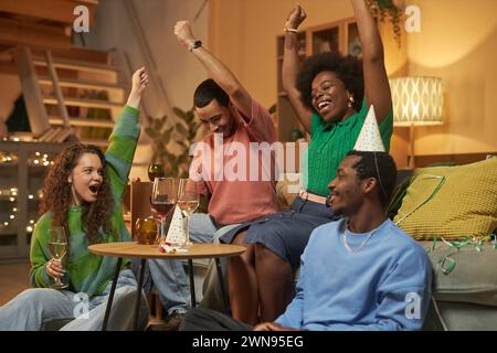 Porträt multiethnischer junger Menschen, die fröhlich jubeln und die Hände heben, um den Sieg zu feiern, während sie gemeinsam während einer gemütlichen Party zu Hause Spiele spielen Stockfoto