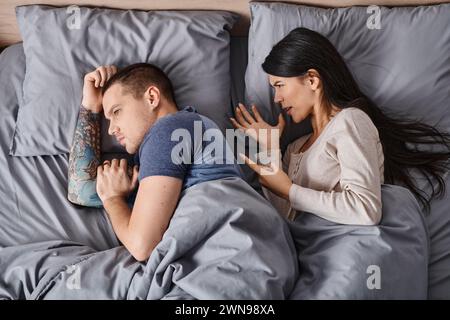 Blick von oben auf eine wütende asiatische Frau, die sich um einen tätowierten jungen Mann streitet, der auf dem Bett lag, Scheidung Stockfoto