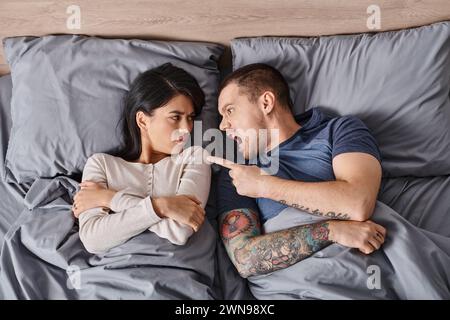Blick von oben auf irritierte interrassische Paare, die auf dem Bett liegen und sich gegenseitig anbrüllen und streiten Stockfoto