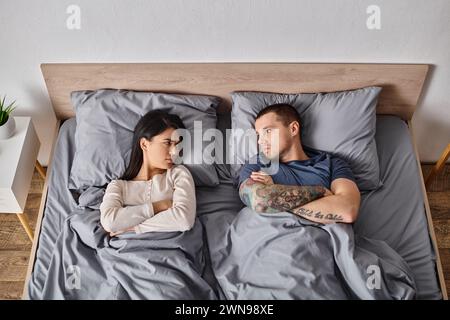 Blick von oben auf beleidigte interrassische Paare, die auf dem Bett liegen und sich gegenseitig ansehen, Konflikt Stockfoto