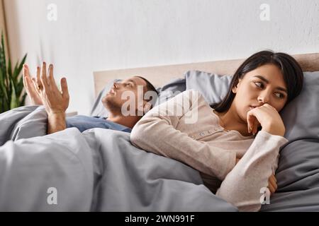 Junge depressive asiatische Frau, die auf dem Bett liegt, nahe dem streitenden Ehemann, Beziehungskonflikt Stockfoto