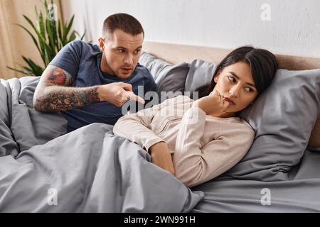Wütender tätowierter Mann, der auf frustrierte asiatische Frau zu Hause im Bett zeigt und streitet, Scheidung Stockfoto