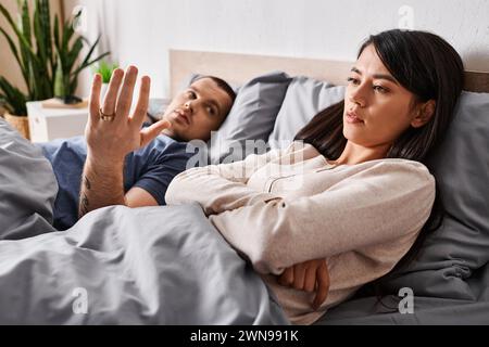 Junger tätowierter Mann streitet um eine verärgerte asiatische Frau zu Hause im Bett, Beziehungsschwierigkeiten Stockfoto