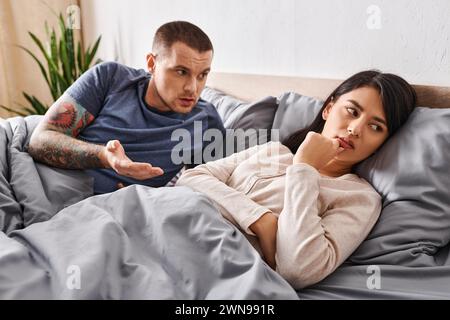 Irritierter tätowierter Mann streitet um verärgerte asiatische Frau zu Hause im Bett, familiäre Schwierigkeiten Stockfoto