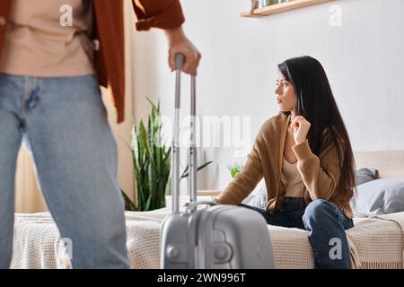 Junge und traurige asiatische Frau, die auf dem Bett sitzt, während ihr Mann mit Koffer das Haus verlässt, Scheidung Stockfoto