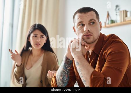 Frustrierte asiatische Frau streitet über verärgerten tätowierten Ehemann zu Hause, familiäres Beziehungsproblem Stockfoto