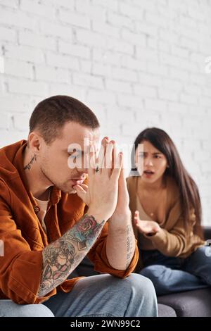 Junger tätowierter Mann, der mit geschlossenen Augen neben einem wütenden asiatischen Mann sitzt, der zu Hause streitet, Scheidung Stockfoto