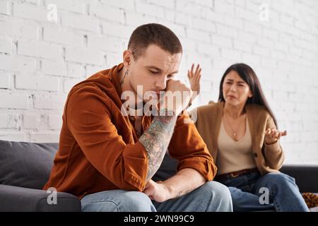 Irritierte asiatische Frau streitet über verärgerten tätowierten Ehemann zu Hause auf der Couch, Familienproblem Stockfoto