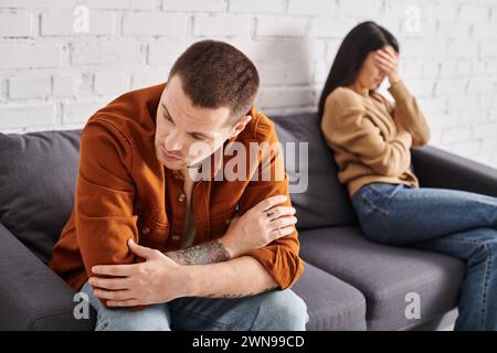 Verärgerter tätowierter Mann, der auf der Couch im Wohnzimmer sitzt, neben weinender asiatischer Frau, Scheidungskonzept Stockfoto