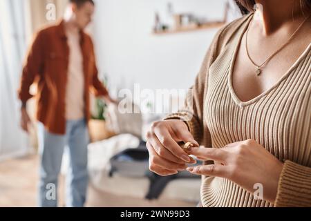 Zugeschnittene Ansicht einer Frau, die den Ehering in der Nähe des Ehemanns abreißt, Koffer im Schlafzimmer packt, Scheidung Stockfoto