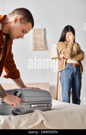 Junger Mann, der Koffer in der Nähe einer depressiven asiatischen Frau packt, die im Schlafzimmer zu Hause weint, Familie scheidet Stockfoto