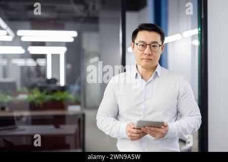 Porträt eines seriösen und selbstbewussten asiatischen Mannes, der in einem weißen Hemd und einer Brille im Büro steht, ein Tablet hält und in die Kamera blickt. Stockfoto