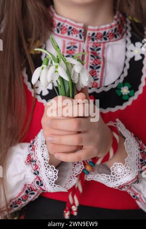 Bulgarisches Mädchen in traditionellen Folklore-Kostümen mit Frühlingsblumen, Schneetropfen und handgefertigtem Wollarmband Martenitsa Symbol von Baba Marta Stockfoto