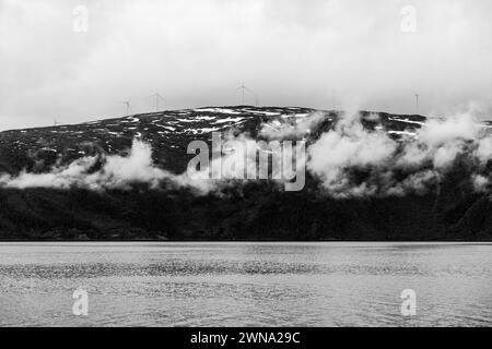 Starke schwarz-weiße Zusammensetzung von Windturbinen auf einem zerklüfteten, schneebedeckten Berg mit Wolkenformationen, die über einem ruhigen See schweben Stockfoto