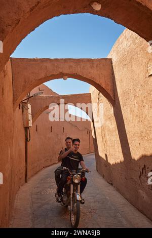 Zwei junge iranische Männer lächeln und zeigen die Daumen hoch, während sie auf dem Motorrad in der engen Straße im historischen Fahadan-Viertel Yazd, Iran, fahren. Stockfoto