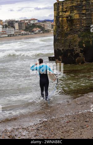 Ein Surfer wandert in Richtung der Wellen, Surfbrett in der Hand, vor der Kulisse der städtischen Küste, bereit, das Meer zu genießen. Stockfoto