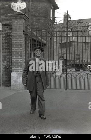 1950er Jahre, historisches Foto, das Bill Baker zeigt, einen langjährigen Mitarbeiter mit 60 Jahren treuer Arbeit, der an den Werkstoren seines ehemaligen Arbeitgebers Lucys, Oxford, England, Großbritannien, stand. Das Geschäft von W. Lucy & Co stammt aus dem Jahr 1812, als William Carter ein Eisenwarengeschäft an der Oxford High Street eröffnete. 1825 zog es in eine neue Messing- und Eisengießerei in Jericho, Oxford, die später als Eagle Works bekannt war. Stockfoto