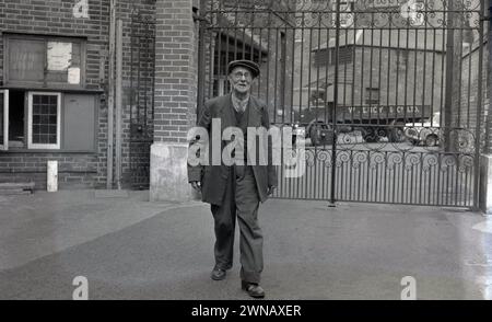 1950er Jahre, historisches Foto, das Bill Baker zeigt, einen langjährigen Mitarbeiter mit 60 Jahren treuer Arbeit, der an den Werkstoren seines ehemaligen Arbeitgebers Lucys, Oxford, England, Großbritannien, stand. Das Geschäft von W. Lucy & Co stammt aus dem Jahr 1812, als William Carter ein Eisenwarengeschäft an der Oxford High Street eröffnete. 1825 zog es in eine neue Messing- und Eisengießerei in Jericho, Oxford, die später als Eagle Works bekannt war. Stockfoto