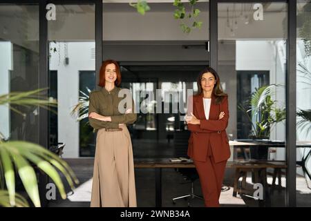 Zwei lächelnde, selbstbewusste Damen, die Arme im Büro kreuzen, Porträt. Stockfoto