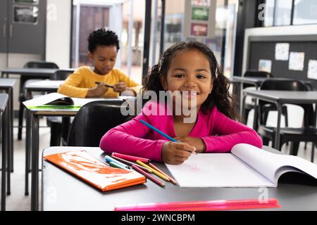 Ein birassisches Mädchen mit lockigen Haaren lächelt beim Zeichnen in einem Schulzimmer, trägt ein pinkfarbenes Oberteil Stockfoto