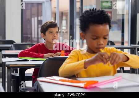 Birassischer Junge in Rot sieht neugierig in der Schulklasse aus, afroamerikanischer Junge konzentriert sich auf das Schreiben Stockfoto