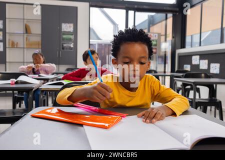 Biracial Boy in Gelb konzentriert sich auf das Schreiben in einem Klassenzimmer; Klassenkameraden arbeiten im Hintergrund Stockfoto