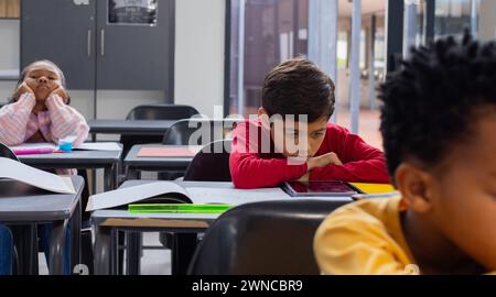 Birassischer Junge in Rot sieht traurig an seinem Schreibtisch in einem Schulzimmer aus, mit Kopierraum, Klassenkameraden herum Stockfoto