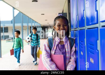 Ein birassisches Mädchen mit einem lila Rucksack lächelt an den blauen Schließfächern der Schule, Jungs im Hintergrund Stockfoto